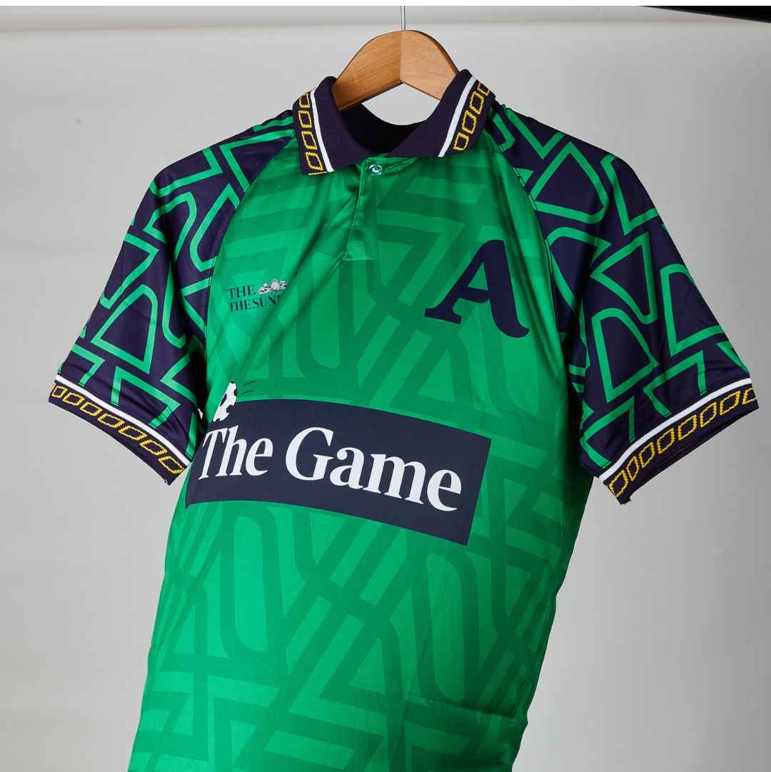 A green and navy blue patterned short-sleeve rugby jersey hanging on a wooden hanger against a light gray background. The jersey features 'The Game' sponsor logo across the chest, a large letter 'A' on the right chest, and an 'Acast' logo near the bottom hem.