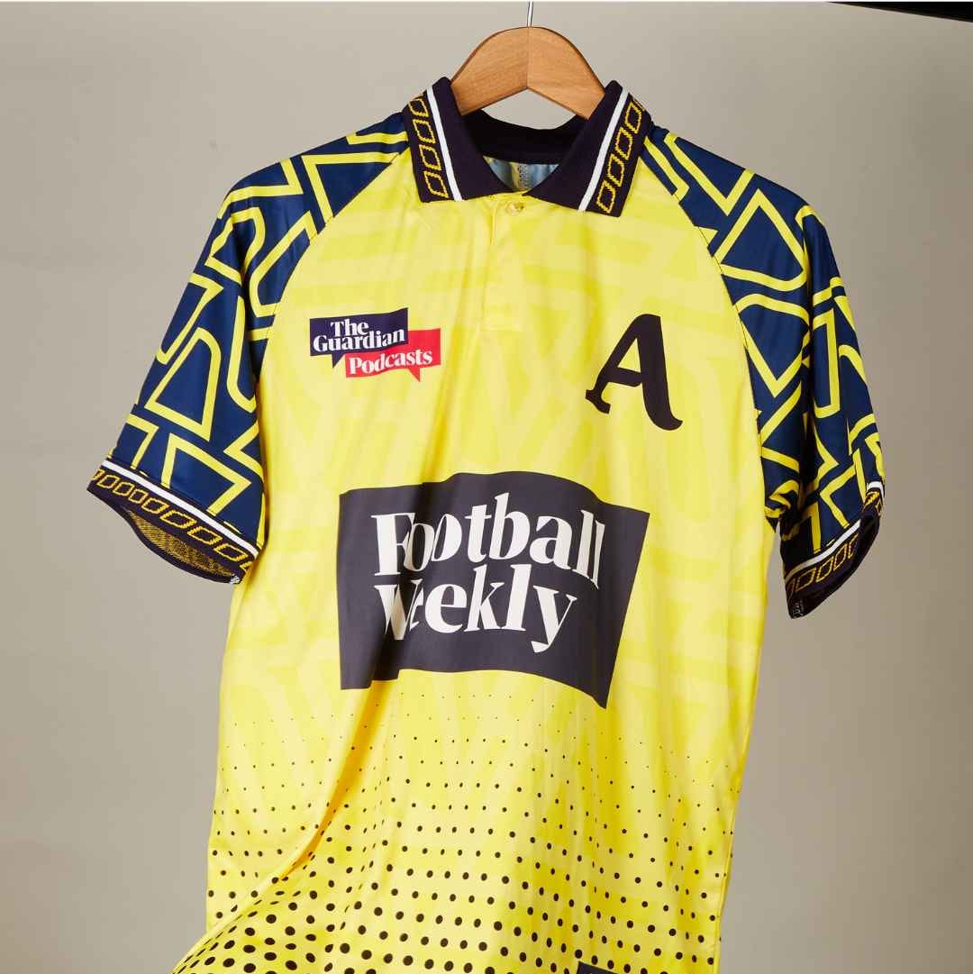 A vintage-style football jersey hanging on a wooden hanger against a plain indoor wall. The shirt is predominantly bright yellow with navy blue geometric patterned sleeves and hem, black dotted gradient at the bottom, and large sponsor logos including Football Weekly and a G4100 mark.